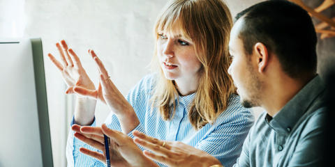 Image shows two coworkers collaborating in front of a computer screen. One coworker is explaining a concept to the other.