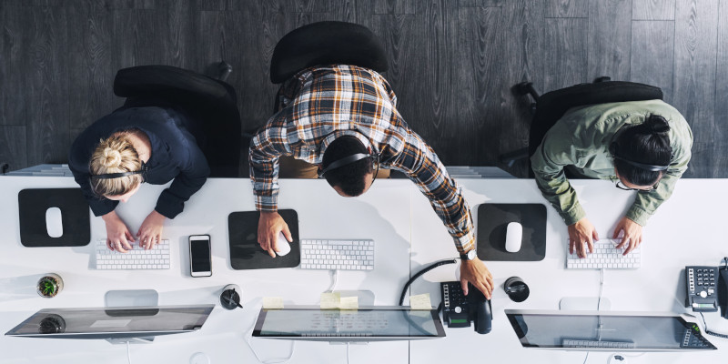 An overhead shot features three teammates working at computers in an office environment.