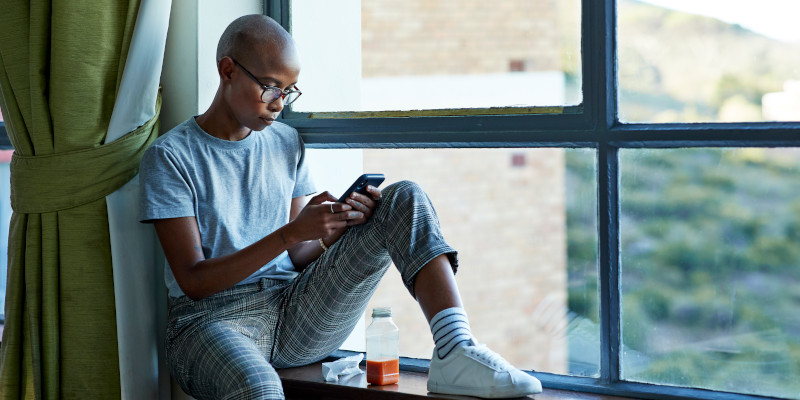 A woman sits in a window sill and glances down at her cell phone.