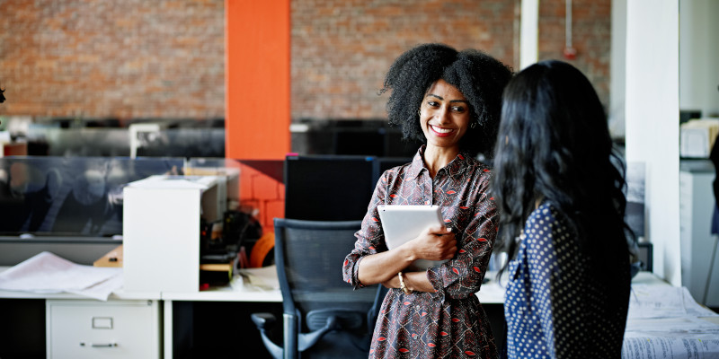 Two women are featured talking in an office environment.