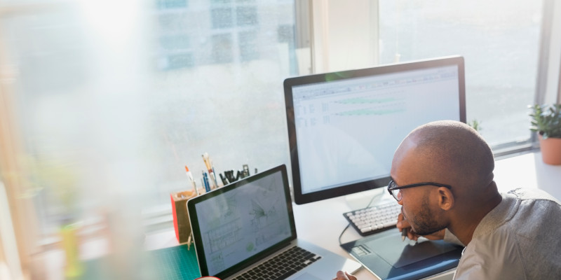 Image shows a man wearing glasses, seated at a desk while working on a laptop. A monitor is pictured in the background.