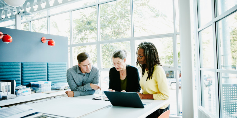 Image of three team members gathered around a table, looking at a laptop.