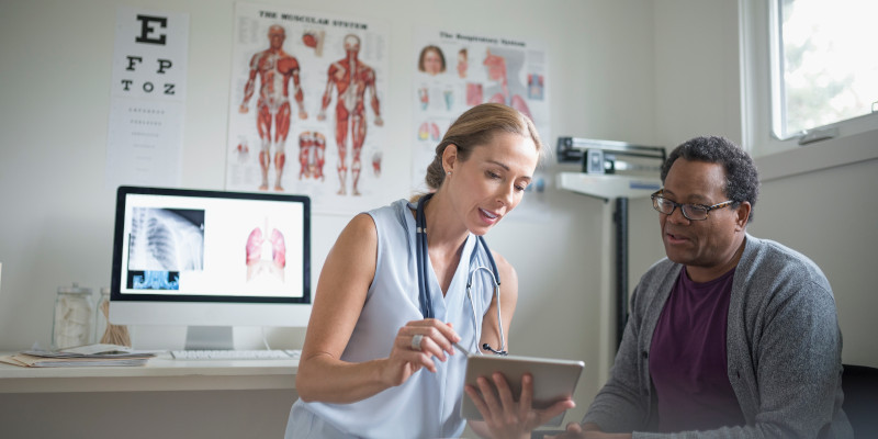 Image shows a doctor speaking to a patient in a hospital setting while holding a tablet.