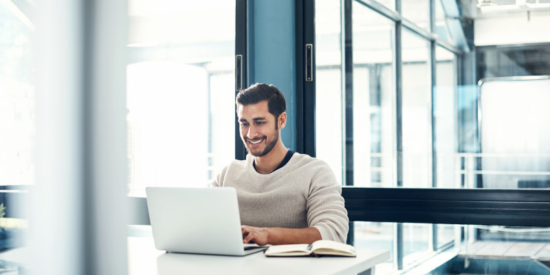 Image shows a man seated at a desk working on a laptop. A book is nearby.