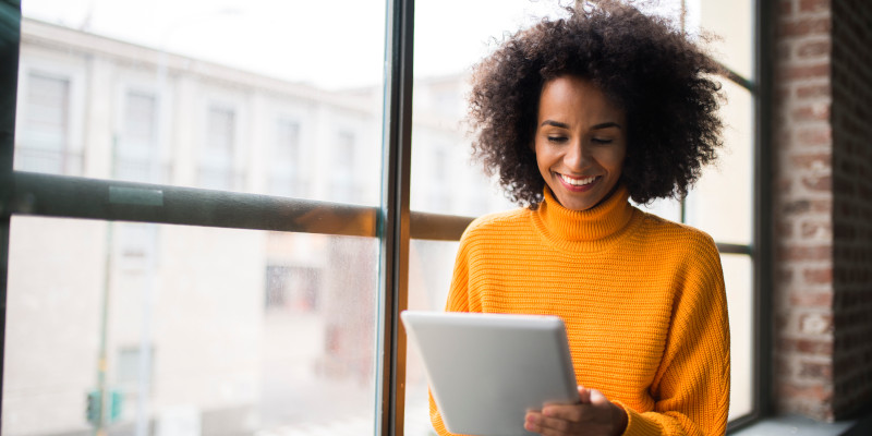 A woman sits by a window and uses a tablet. 